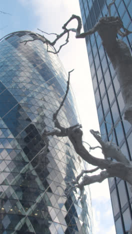 Video-Vertical-Que-Muestra-El-Exterior-Del-Moderno-Edificio-De-Oficinas-Gherkin-En-La-Ciudad-De-Londres,-Reino-Unido,-Con-Escultura-En-Primer-Plano